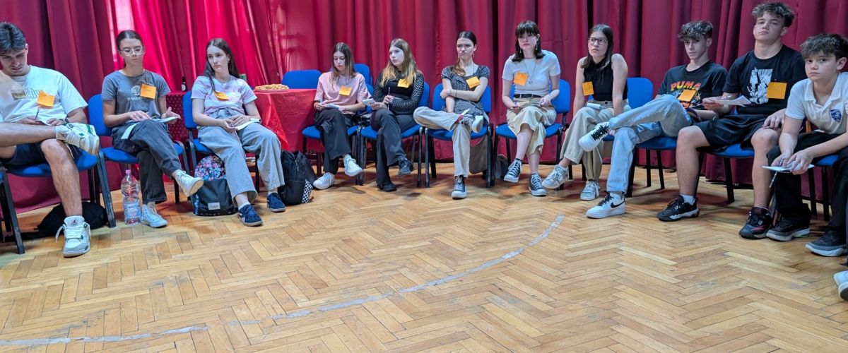 Group of young people seated in a circle in a room with red stage curtains, sharing snacks and chatting during a workshop.
